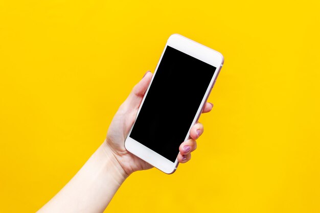 Téléphone blanc avec écran noir dans une main féminine isolée sur un mur jaune de couleur vive