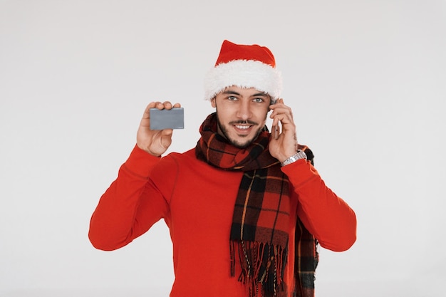 Avec téléphone et carte de crédit Jeune bel homme en vêtements de nouvel an debout à l'intérieur sur fond blanc