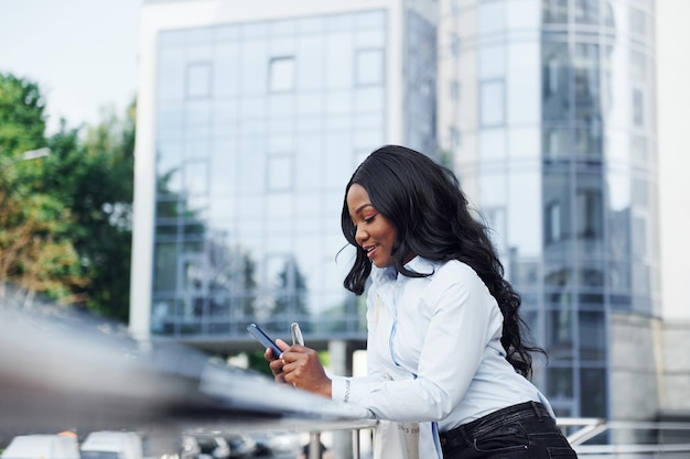 Avec le téléphone dans les mains Jeune femme afro-américaine en chemise blanche à l'extérieur de la ville contre le bâtiment de l'entreprise