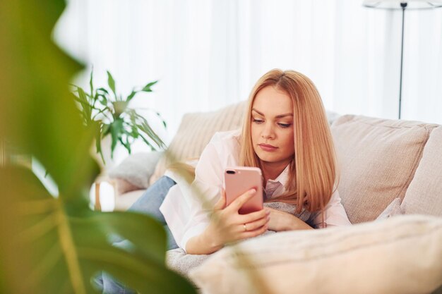 Avec le téléphone dans les mains Jeune femme en chemise blanche et jeans est à la maison