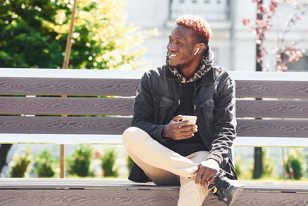 Avec téléphone à la main Jeune homme afro-américain en veste noire à l'extérieur dans la ville assis sur un banc