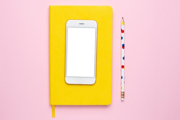 Téléphone mobile avec un cahier jaune et un crayon coloré sur une vue de dessus de fond rose