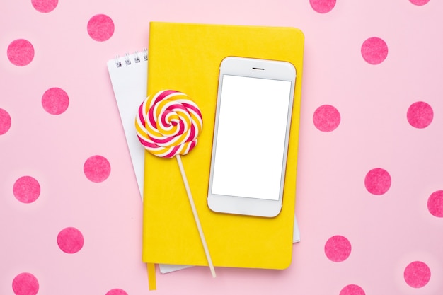 Téléphone mobile avec un cahier jaune et lolipop coloré sur fond rose avec vue de dessus de confettis