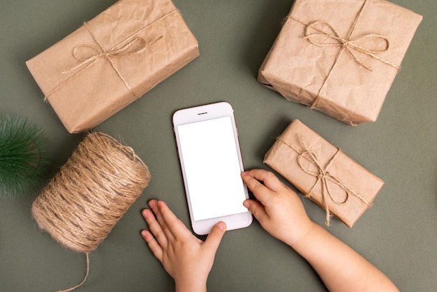 Téléphone mobile de fond de technologie de vacances de Noël dans des mains d'enfant et des boîtes-cadeaux enveloppées de papier d'artisanat ...