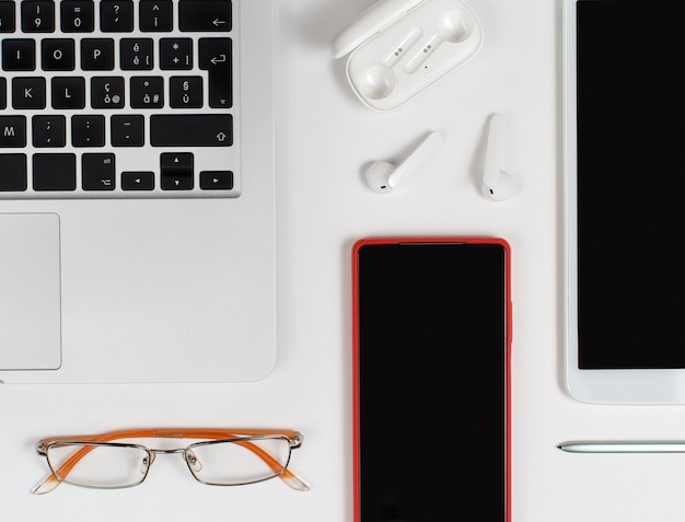 Téléphone mobile rouge, écouteurs et lunettes près d'un ordinateur portable sur fond blanc