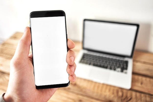 Téléphone portable avec un écran vierge dans la paume de la main masculine, en arrière-plan un ordinateur portable sur une table en bois et sur un fond blanc