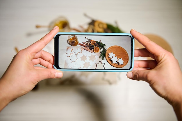 Téléphonez en temps réel. Fermer. Mains tenant le téléphone et prenant des photos de pains d'épice en forme d'étoiles disposées sur une planche de bois et sur du papier sulfurisé à côté de tranches d'orange séchées, de pommes de pin, de brindilles