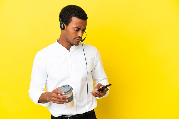 Télévendeur afro-américain travaillant avec un casque sur fond jaune isolé tenant du café à emporter et un mobile