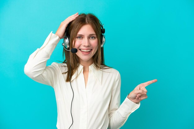 Télévendeur anglais femme travaillant avec un casque isolé sur fond bleu surpris et pointant le doigt sur le côté