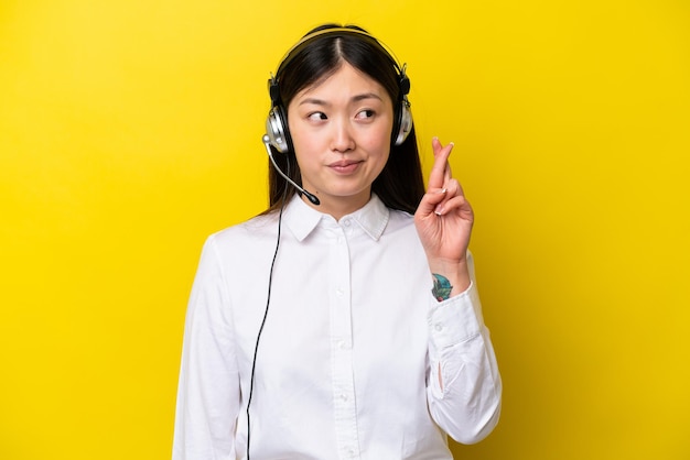 Télévendeur femme chinoise travaillant avec un casque isolé sur fond jaune avec les doigts croisés et souhaitant le meilleur