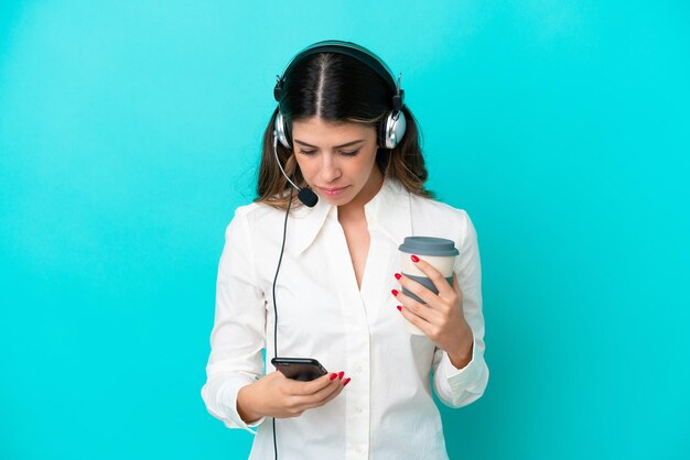 Télévendeur femme italienne travaillant avec un casque isolé sur fond bleu tenant du café à emporter et un mobile