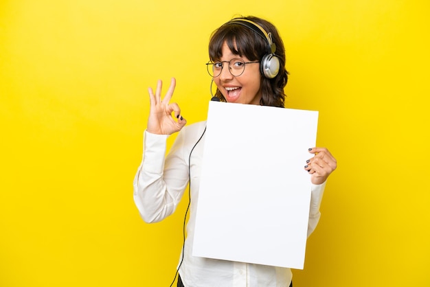 Télévendeur femme latine travaillant avec un casque isolé sur fond jaune tenant une pancarte vide et faisant signe OK