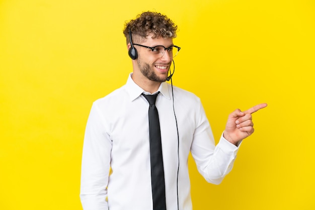 Télévendeur homme blond travaillant avec un casque isolé sur fond jaune pointant le doigt sur le côté et présentant un produit