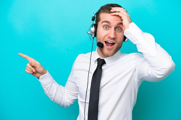 Télévendeur homme caucasien travaillant avec un casque isolé sur fond bleu surpris et pointant le doigt sur le côté