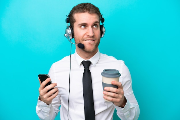 Télévendeur homme caucasien travaillant avec un casque isolé sur fond bleu tenant du café à emporter et un mobile tout en pensant à quelque chose