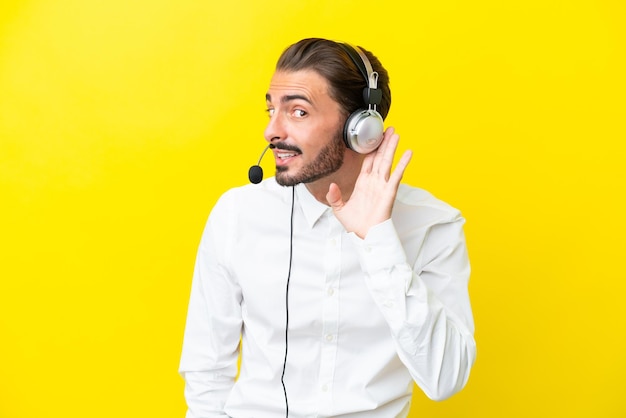 Télévendeur homme caucasien travaillant avec un casque isolé sur fond jaune écoutant quelque chose en mettant la main sur l'oreille