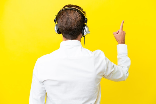 Télévendeur homme caucasien travaillant avec un casque isolé sur fond jaune pointant vers l'arrière avec l'index