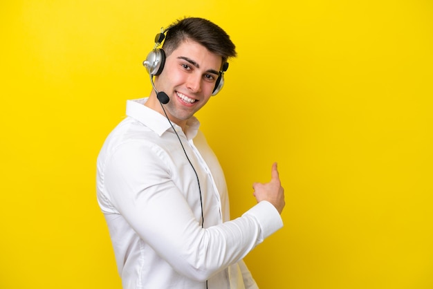 Télévendeur homme caucasien travaillant avec un casque isolé sur fond jaune pointant vers l'arrière