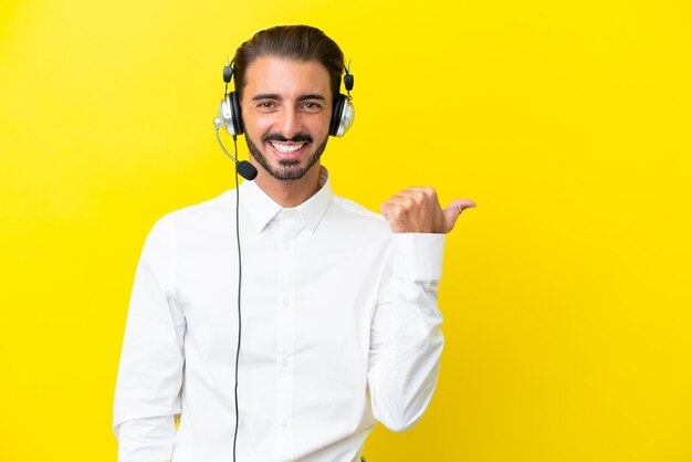 Télévendeur homme caucasien travaillant avec un casque isolé sur fond jaune pointant vers le côté pour présenter un produit