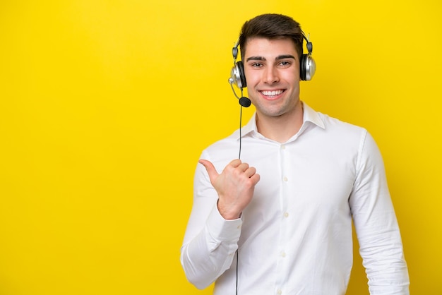 Télévendeur homme caucasien travaillant avec un casque isolé sur fond jaune pointant vers le côté pour présenter un produit