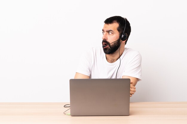 Télévendeur homme caucasien travaillant avec un casque et avec un ordinateur portable faisant un geste surprise tout en regardant sur le côté