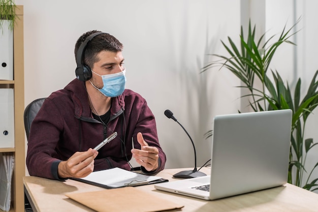 Télévendeur ou homme podcast travaillant avec un ordinateur portable et parlant sur un casque avec masque facial au bureau