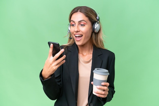 Télévendeur jolie femme uruguayenne travaillant avec un casque sur fond isolé tenant du café à emporter et un mobile