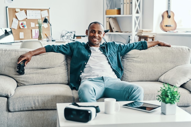 Tellement bon d'être à la maison! Beau jeune homme africain regardant la caméra et souriant alors qu'il était assis à l'intérieur