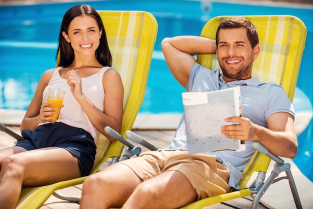 Tellement bon de ne rien faire ! Heureux jeune couple se détendre ensemble assis sur les transats au bord de la piscine