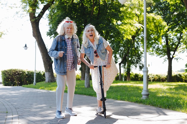 Tellement chouette. Joyeuse femme heureuse à l'aide d'un scooter tout en étant dans le parc avec son mari