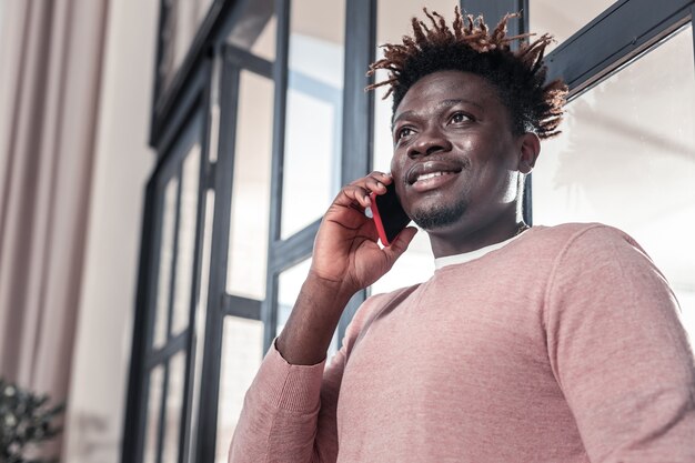 Tellement heureux. Bel homme international parlant par téléphone et debout près de la fenêtre