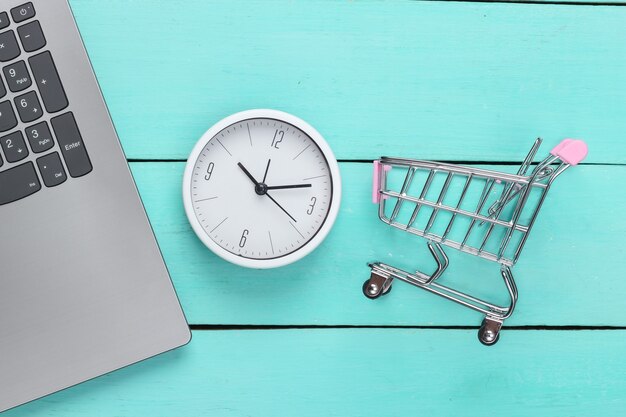 Temps d'achats en ligne. Clavier d'ordinateur avec horloge et chariot de supermarché sur une surface en bois bleue. Vue de dessus