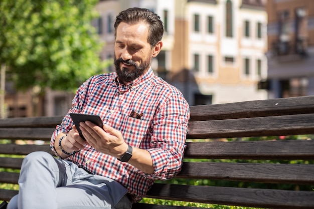 Temps agréable. Heureux homme adulte lisant son livre électronique tout en se reposant sur le banc