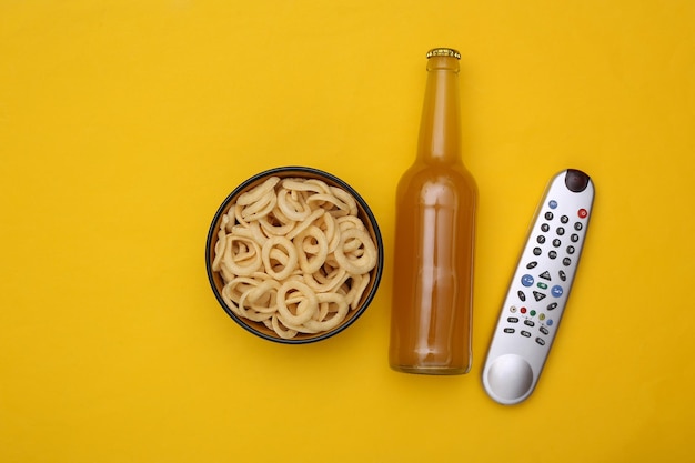 Le temps du film. Bouteille de bière et bol avec collation aux anneaux de maïs, télécommande tv sur fond noir. Vue de dessus