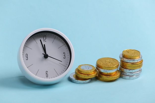 Le temps, c'est de l'argent. Horloge blanche avec une pile de pièces sur un bleu