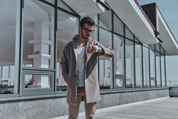 Le temps, c'est de l'argent. Jeune homme confiant en lunettes regardant sa montre en se tenant debout à l'extérieur