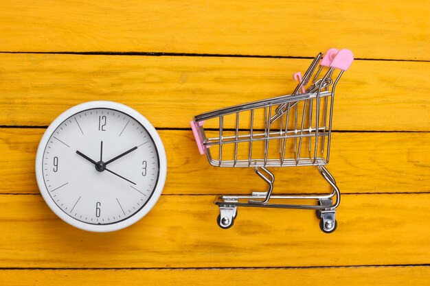 Temps de magasinage. Chariot de supermarché avec horloge sur une surface en bois jaune. Minimalisme. Vue de dessus