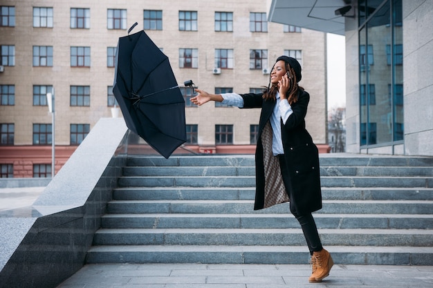 Temps orageux. Une fille noire attrape un parapluie volant en parlant au téléphone portable dans la rue, concept de légèreté