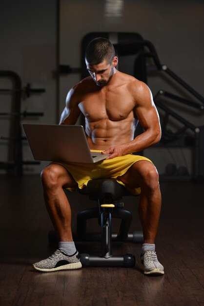 Temps de repos Jeune homme musclé confiant se reposant dans une salle de sport saine et utilisant un ordinateur portable