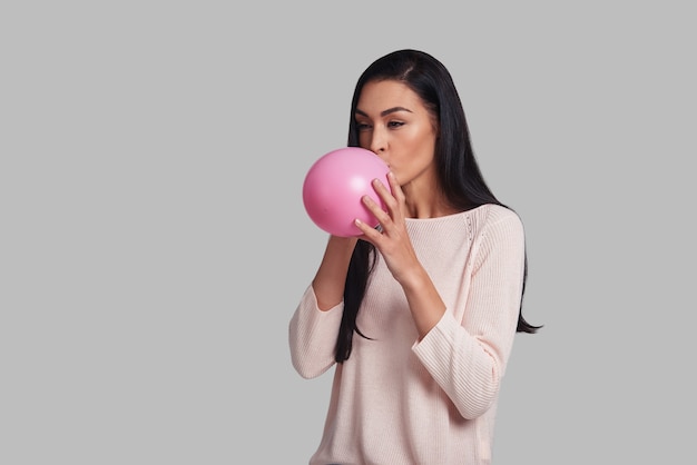 Le temps de s'amuser ! Prise de vue en studio d'une jeune femme séduisante en tenue décontractée faisant exploser un ballon rose en se tenant debout sur fond gris