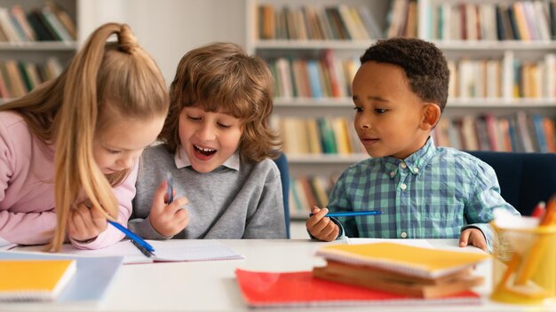 Temps scolaire Enfants multiethniques écrivant dans des cahiers et souriant, parlant entre eux et riant assis à table