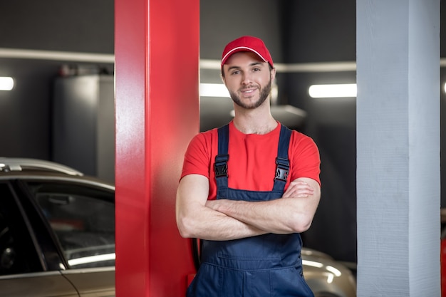 Temps de travail. Souriant jeune homme barbu en salopette de travail et casquette debout dans l'atelier automobile