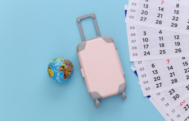 Le temps de voyager. Mini valise de voyage en plastique, globe et calendrier mensuel sur fond bleu. Style minimaliste. Vue de dessus. Mise à plat