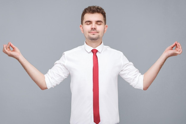 Temps de yoga. Portrait de jeune bel homme calme en chemise blanche et cravate debout dans la pose de yoga et essayez de vous détendre. intérieur isolé sur fond gris.