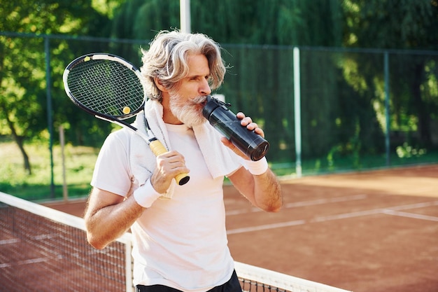 Tenant une bouteille d'eau Senior homme élégant moderne avec une raquette à l'extérieur sur un court de tennis pendant la journée