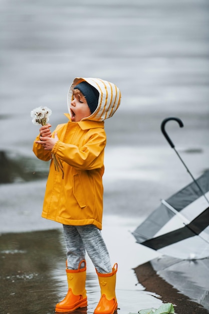 Tenant des fleurs Enfant dans des bottes imperméables jaunes et avec un parapluie jouant à l'extérieur après la pluie