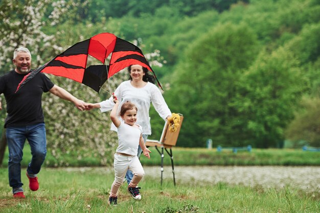 Tenant par une main. Enfant de sexe féminin positif avec grand-mère et grand-père en cours d'exécution avec cerf-volant de couleur rouge et noir dans les mains à l'extérieur