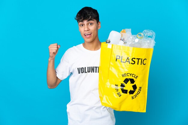 Tenant un sac plein de bouteilles en plastique à recycler sur bleu célébrant une victoire en position de vainqueur