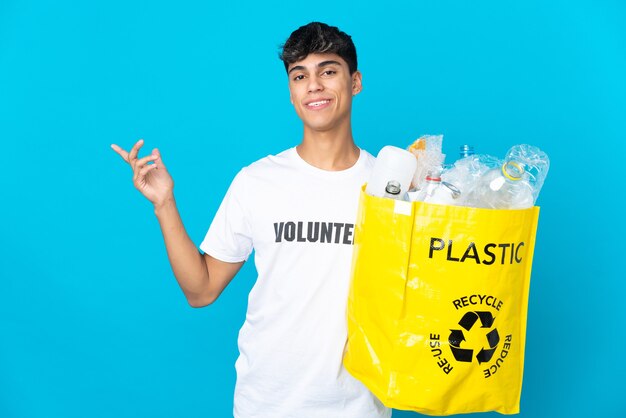 Tenant un sac plein de bouteilles en plastique à recycler sur un mur bleu en étendant les mains sur le côté pour inviter à venir