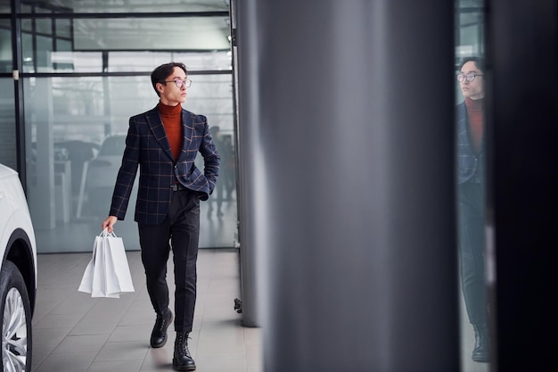 Tenant des sacs à provisions. Jeune homme d'affaires en costume de luxe et vêtements formels est à l'intérieur près de la voiture.